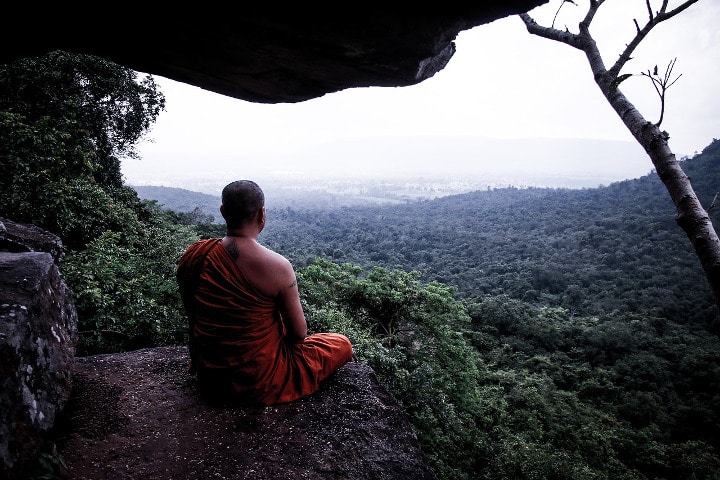 monk meditating outside of cave entrance overlooking a forest
